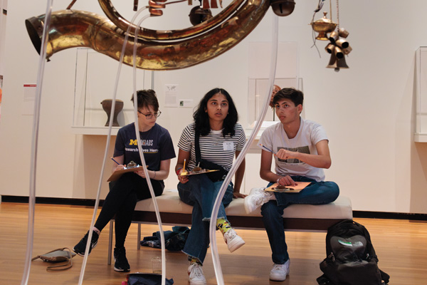 Students discussing a suspended sculpture of musical instruments in UMMA